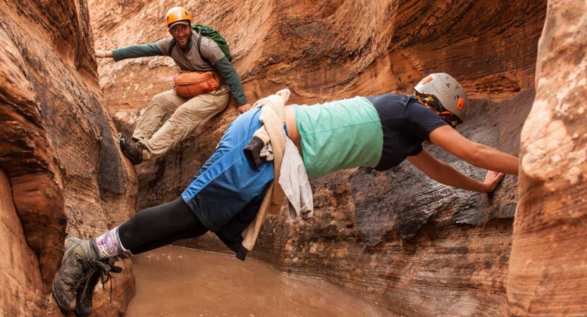 two people wearing helmets make their way through a water-filled slot canyon by using their arms and legs on either side of the walls